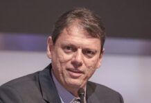 Um homem de pele clara e cabelos curtos castanhos aparece em um plano fechado, falando em um microfone que está posicionado logo abaixo de seu rosto. Ele veste um paletó escuro sobre uma camisa social azul clara e gravata estampada. Sua expressão é séria e focada. O fundo está desfocado em tons de cinza e lilás.