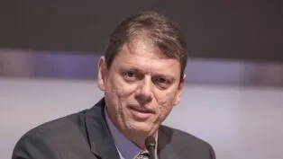 Um homem de pele clara e cabelos curtos castanhos aparece em um plano fechado, falando em um microfone que está posicionado logo abaixo de seu rosto. Ele veste um paletó escuro sobre uma camisa social azul clara e gravata estampada. Sua expressão é séria e focada. O fundo está desfocado em tons de cinza e lilás.