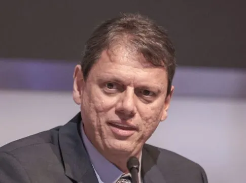 Um homem de pele clara e cabelos curtos castanhos aparece em um plano fechado, falando em um microfone que está posicionado logo abaixo de seu rosto. Ele veste um paletó escuro sobre uma camisa social azul clara e gravata estampada. Sua expressão é séria e focada. O fundo está desfocado em tons de cinza e lilás.