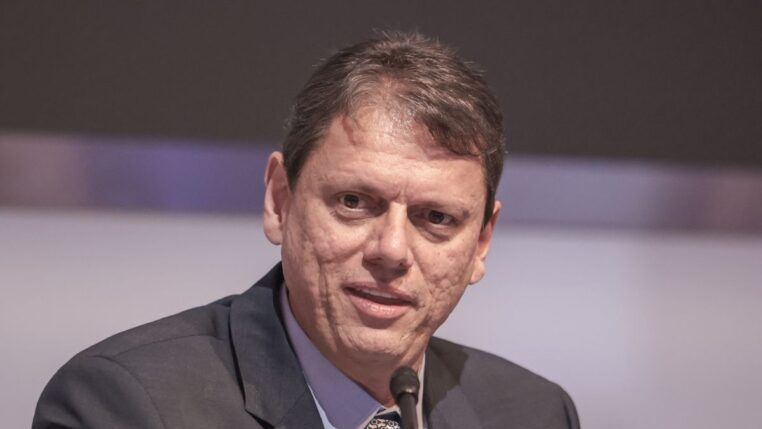 Um homem de pele clara e cabelos curtos castanhos aparece em um plano fechado, falando em um microfone que está posicionado logo abaixo de seu rosto. Ele veste um paletó escuro sobre uma camisa social azul clara e gravata estampada. Sua expressão é séria e focada. O fundo está desfocado em tons de cinza e lilás.