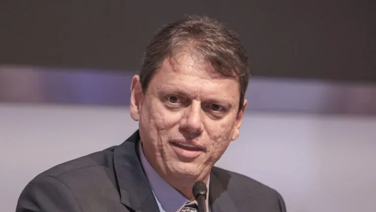 Um homem de pele clara e cabelos curtos castanhos aparece em um plano fechado, falando em um microfone que está posicionado logo abaixo de seu rosto. Ele veste um paletó escuro sobre uma camisa social azul clara e gravata estampada. Sua expressão é séria e focada. O fundo está desfocado em tons de cinza e lilás.