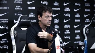 O técnico Fernando Diniz concede uma entrevista coletiva. Ele está sentado em uma cadeira esportiva preta e branca com o escudo do Vasco da Gama, vestindo uma camisa polo preta. Com a mão direita levemente levantada em um gesto explicativo, ele olha para a frente. À sua frente, sobre a mesa, há um microfone com a marca "VascoTV", uma pequena bandeira do clube, um tablet e copos personalizados. O fundo é um painel preto repleto de logotipos de patrocinadores como "Pixbet", "Brahma", "Bmg" e o símbolo da Nike.