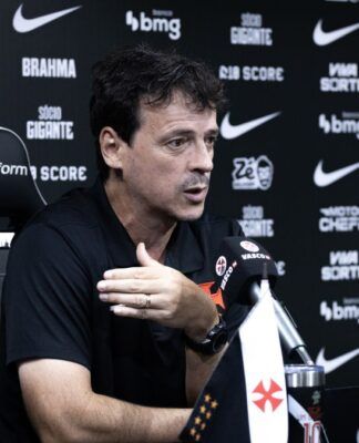 O técnico Fernando Diniz concede uma entrevista coletiva. Ele está sentado em uma cadeira esportiva preta e branca com o escudo do Vasco da Gama, vestindo uma camisa polo preta. Com a mão direita levemente levantada em um gesto explicativo, ele olha para a frente. À sua frente, sobre a mesa, há um microfone com a marca 