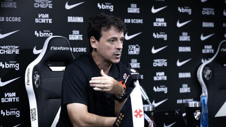 O técnico Fernando Diniz concede uma entrevista coletiva. Ele está sentado em uma cadeira esportiva preta e branca com o escudo do Vasco da Gama, vestindo uma camisa polo preta. Com a mão direita levemente levantada em um gesto explicativo, ele olha para a frente. À sua frente, sobre a mesa, há um microfone com a marca "VascoTV", uma pequena bandeira do clube, um tablet e copos personalizados. O fundo é um painel preto repleto de logotipos de patrocinadores como "Pixbet", "Brahma", "Bmg" e o símbolo da Nike.
