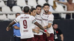 Quatro jogadores de futebol do Fluminense FC celebram juntos no gramado. Três deles estão de frente, abraçados, vestindo a camisa branca com detalhes em grená e verde e calções grenás. O quarto jogador está de costas, exibindo o nome "JOHN KENNEDY" e o número 9 na camisa. Ao fundo, as arquibancadas do estádio mostram cadeiras brancas e pretas, com alguns torcedores e profissionais de imprensa visíveis de forma desfocada.