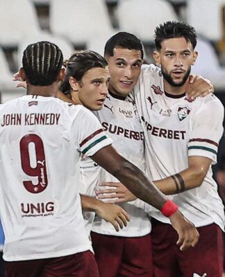 Quatro jogadores de futebol do Fluminense FC celebram juntos no gramado. Três deles estão de frente, abraçados, vestindo a camisa branca com detalhes em grená e verde e calções grenás. O quarto jogador está de costas, exibindo o nome 
