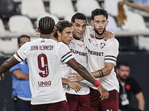 Quatro jogadores de futebol do Fluminense FC celebram juntos no gramado. Três deles estão de frente, abraçados, vestindo a camisa branca com detalhes em grená e verde e calções grenás. O quarto jogador está de costas, exibindo o nome 