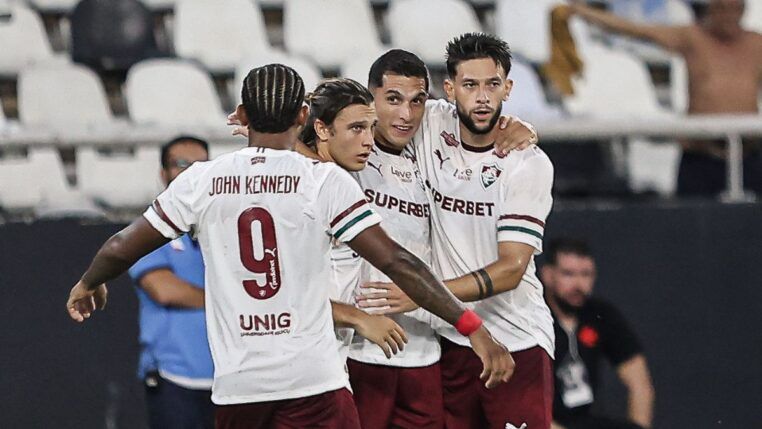 Quatro jogadores de futebol do Fluminense FC celebram juntos no gramado. Três deles estão de frente, abraçados, vestindo a camisa branca com detalhes em grená e verde e calções grenás. O quarto jogador está de costas, exibindo o nome "JOHN KENNEDY" e o número 9 na camisa. Ao fundo, as arquibancadas do estádio mostram cadeiras brancas e pretas, com alguns torcedores e profissionais de imprensa visíveis de forma desfocada.
