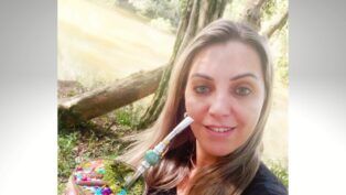 Roseli Vanda Pires Albuquerque tomando chimarrão em espaço público aberto, aparentemente um parque, perto de uma árvore