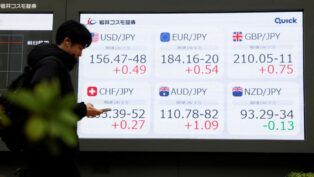 Pessoa olha o celular diante de painel com números do mercado de ações