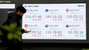 Pessoa olha o celular diante de painel com números do mercado de ações