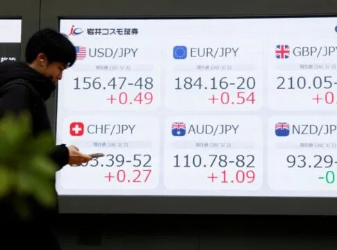 Pessoa olha o celular diante de painel com números do mercado de ações