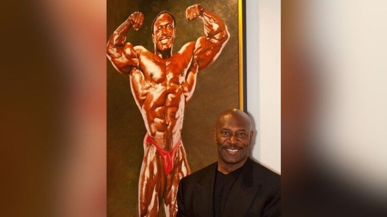 Lee Haney veste terno preto e aparece à frente de uma foto antiga sua, na época de competições