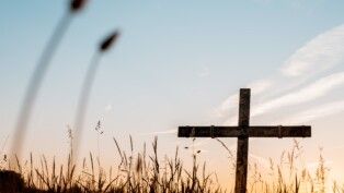 Uma cruz, no fundo um céu azul, fixa em um campo de trigo
