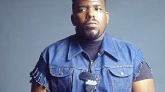 Afrika Bambaataa olhando sério para a câmera. Ele usa um colete jeans.