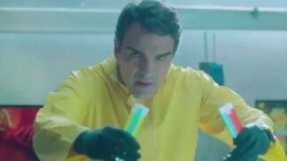 Homem usa uma roupa de laboratório amarela e segura dois frascos com cores diferentes.