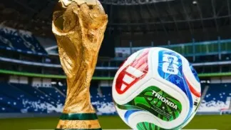 Taça e bola da Copa do Mundo de 2026 em foto, com um estádio no fundo.
