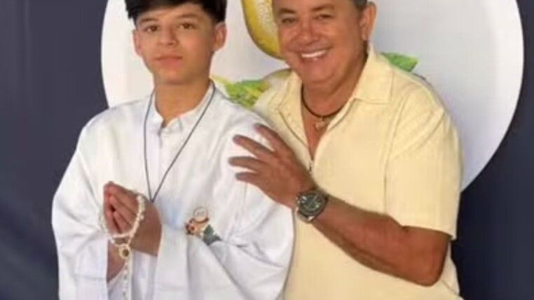 Na foto há um homem e ao seu lado um adolescente com vestimentas comuns da primeira comunhão.