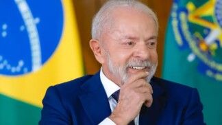 Lula ri para a câmera e atrás dele tem duas bandeiras.