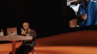 Homem sentado em um palco com vídeos passando atrás dele.