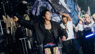 Homem com jaqueta preta e atrás dele outro homem segura uma bandeira.