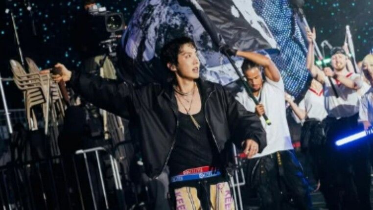 Homem com jaqueta preta e atrás dele outro homem segura uma bandeira.