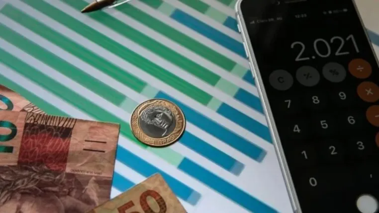 Notas de real, moedas e um celular com aplicativo de calculadora sobre um gráfico