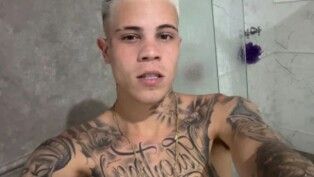 Homem de cabelo platinado e sem camiseta com diversas tatuagens.