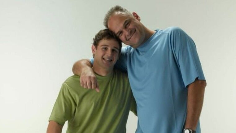 Oscar e seu filho abraçados. Oscar usa blusa azul e seu filho verde.