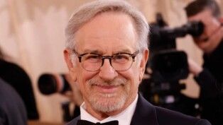 Steven Spilberg sorrindo para a câmera, ele usa terno e óculos.