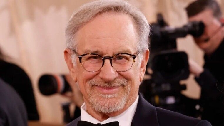 Steven Spilberg sorrindo para a câmera, ele usa terno e óculos.