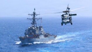 O destróier USS Donald Cook (DDG-75) navega pelo Mar Arábico enquanto um helicóptero MH-60S Sea Hawk sobrevoa a região. As forças dos Estados Unidos afirmam manter o bloqueio naval contra o Irã. Segundo o Comando Central dos EUA (CENTCOM), até 18/07/2026, cinco embarcações comerciais foram redirecionadas e uma foi interceptada.