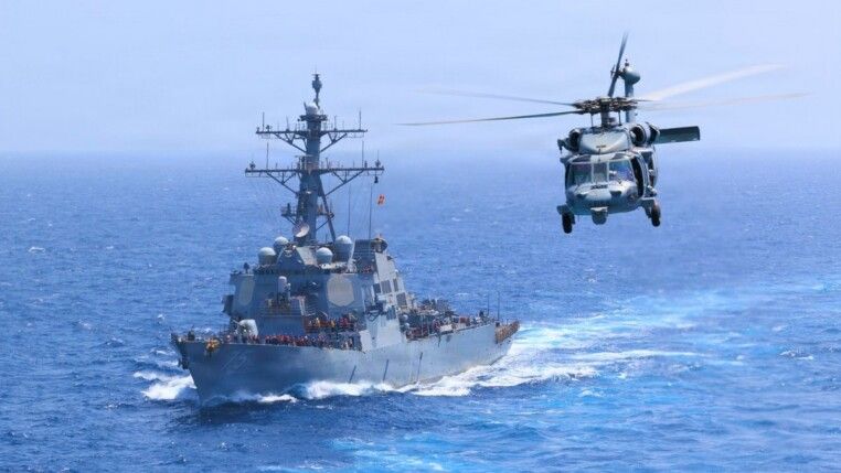 O destróier USS Donald Cook (DDG-75) navega pelo Mar Arábico enquanto um helicóptero MH-60S Sea Hawk sobrevoa a região. As forças dos Estados Unidos afirmam manter o bloqueio naval contra o Irã. Segundo o Comando Central dos EUA (CENTCOM), até 18/07/2026, cinco embarcações comerciais foram redirecionadas e uma foi interceptada.