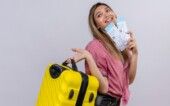 gastos-turismo-viagem Jovem mulher sorridente usando camiseta vermelha segura uma mala amarela e passagens aéreas enquanto olha de lado para uma parede branca.
