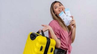 Jovem mulher sorridente usando camiseta vermelha segura uma mala amarela e passagens aéreas enquanto olha de lado para uma parede branca.
