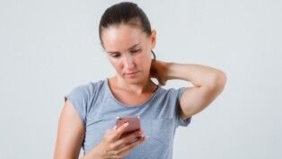 Mulher jovem usando celular com a mão no pescoço, vestindo camiseta cinza, vista frontal.