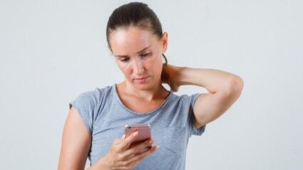Mulher jovem usando celular com a mão no pescoço, vestindo camiseta cinza, vista frontal.