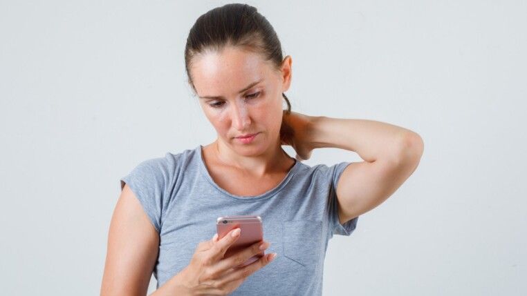 Mulher jovem usando celular com a mão no pescoço, vestindo camiseta cinza, vista frontal.